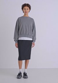 Sweatshirt cinza de manga longa com decote redondo, sobre uma camisa branca, combinado com uma saia lápis preta e ténis pretos.