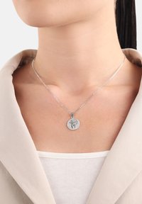 Silverkedjehalsband med ett runt hänge som har en ingraverad design. Hängsmycket är texturerat och hänger framträdande mot en neutral topp.
