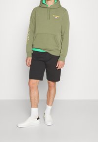 Olivgrön huvtröja med framficka, gul text på ärmarna, svarta shorts, vita slip-on skor och vita strumpor. Enkelt, avslappnat outfit.