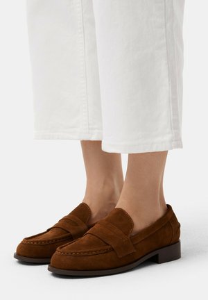 Brązowe zamszowe loafersy z paskiem z przodu, niskim blokowym obcasem i widocznymi detalami szwów, zestawione z krótkimi białymi spodniami.