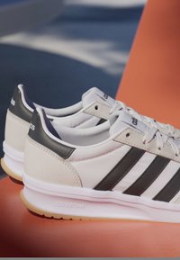 Paar weiße Adidas-Sneaker mit schwarzen Streifen und "Cloudfoam"-Dämpfung, auf einer orangefarbenen Oberfläche mit schattigem Hintergrund.
