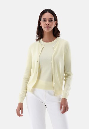Cardigan giallo chiaro con chiusura a bottoni, texture morbida e maniche lunghe, indossato sopra un maglione abbinato e abbinato a pantaloni bianchi.