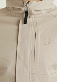 Beige jacka med framdragkedja delvis täckt av en tygflik och en liten broderad geometrisk logotyp på bröstet.