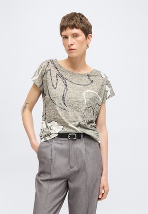 Frau mit kurzen Haaren, die ein grau gemustertes kurzärmeliges Oberteil trägt, das in eine hoch taillierte graue Hose mit schwarzem Gürtel gesteckt ist.