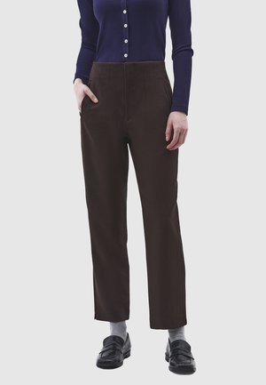 Person iført højtaljede mørkebrune bukser, navyblå langærmet knappet trøje, grå sokker, sorte loafers, med den ene hånd i lommen, stående på grå baggrund.