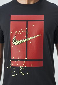 Personne portant un t-shirt noir avec un graphisme de terrain de tennis rouge et des points jaunes formant un logo Nike swoosh sur le design du terrain.