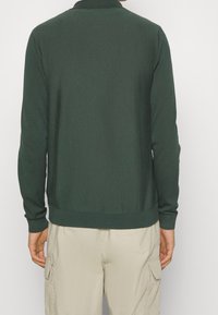 Mörkgrön långärmad tröja med krage, ribbade ärmslut och nederkant. Slät textur, figurnära silhuett och ren design utan mönster.