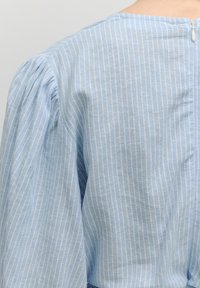 Lichtblauwe blouse met verticale witte strepen, opgeblazen mouwen en een discreet rits aan de achterkant. Zachte textuur en relaxed fit.