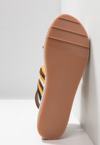 Sandale rayée avec une semelle en caoutchouc, présentant des rayures verticales jaunes, bleu marine et blanches. Accentuée d'une bande marron texturée. Design plat.