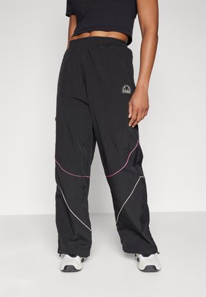 Persona que lleva pantalones negros con cintura elástica con líneas curvas rosas y blancas y zapatillas blancas, de pie frente a un fondo liso.