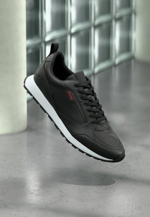 Must nahast tennised valge tallaga ja mustade paeltega, mille küljes on punane "HUGO" logo, hõljuvad hallil poleeritud põrandal.