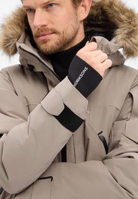 Beige parka met een nepleren capuchon, voorzien van een zwarte verstelbare manchette met logo en klittenbandsluiting. Geweven stof met een standaard pasvorm.
