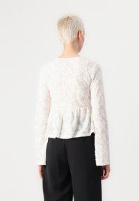 Weiße Spitzenbluse mit langen Ärmeln, aufwendiger floraler Textur, peplumförmigem Saum, rundem Ausschnitt, über schwarzen Hosen getragen. Rückansicht dargestellt.