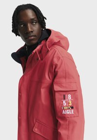 Tamsaus odos žmogus su trumpais dredais, vilkintis koralų raudoną džemperį su kapišonu, kuriame šonuose yra išsiuvinėti skaičiai ir užrašas "AIGLE" ant kišenės.