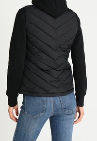 Eine Person trägt eine schwarze gesteppte Weste mit einem Chevron-Muster über einem schwarzen Hoodie und blauen Jeans, mit dem Rücken zur Kamera gewandt.