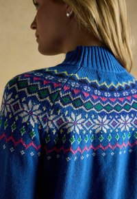 Zils adīts džemperis ar augstu ribotu apkakli, sarežģītām sniega kule un ģeometriskām rakstām baltā, rozā, zaļā un dzeltenā krāsā.