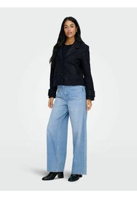 Veste noire texturée coupée avec un col à rabat, deux boutons et des poches avant, associée à un jeans taille haute bleu clair à jambes larges et des bottines noires.