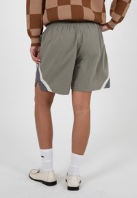 Personne portant un short vert avec des panneaux latéraux blancs et gris, des chaussettes blanches, des mocassins crème et un pull marron à carreaux, vue de dos.