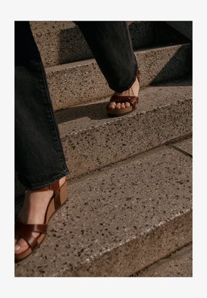 Sandales à plateforme marron avec des brides croisées, dotées d'un talon en liège texturé, portées sur un pied montant des escaliers en pierre, associées à un jean foncé.