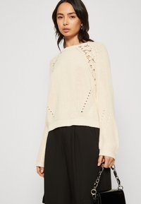 Creme strikket sweater med åbent arbejdet detaljering ved skuldrene, med en afslappet pasform og ribbede manchetter, parret med sorte vide bukser.