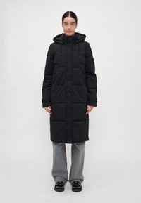 Crni puffer kaput s kapuljačom, dugog dizajna, s podesivim šnurama i quilted teksturom. Nošen preko traperica i tenisica.