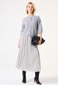Femme portant un cardigan gris boutonné, une jupe midi plissée argentée, des bottes à talons noires, tenant une grande pochette noire contre un fond neutre.