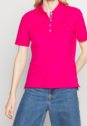 Polo fuchsia à manches courtes, col ouvert, patte de boutonnage à deux boutons et un petit logo sur la poitrine. Associé à un jean en denim bleu.