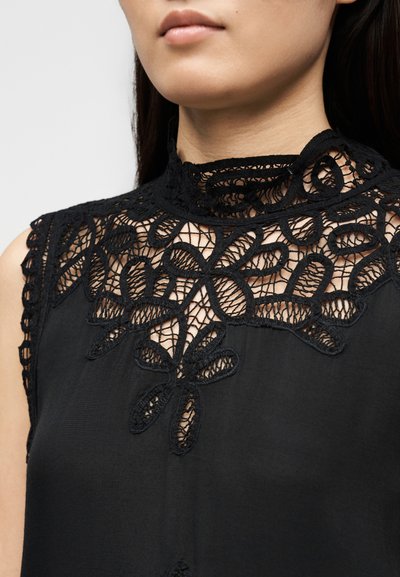 Γυναίκα που φοράει αμάνικη μαύρη μπλούζα με ψηλό δαντελένιο γιακά και φλοράλ δαντελένιο σχέδιο στο πάνω μέρος του στήθους.