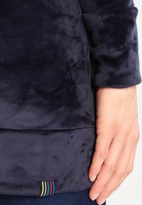 Mano appoggiata accanto all'orlo di una giacca in velluto blu scuro con un'etichetta a strisce multicolori vicino al bordo inferiore.