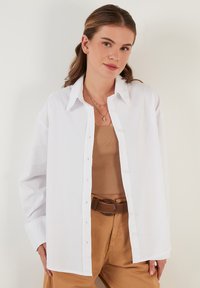 White button-up shirt made of lightweight fabric, featuring long sleeves and a structured collar, paired with tan high-waisted trousers.