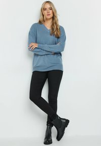 Helle blaue Übergröße-Pullover mit V-Ausschnitt, kombiniert mit schwarzen Slim-Fit-Hosen und schwarzen Knöchelstiefeln mit strukturierten Sohlen.