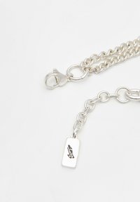 Strieborný reťazový náhrdelník s karabínkovým uzáverom a malou obdĺžnikovou štítkom s vygravírovaným podpisom na bielom pozadí.