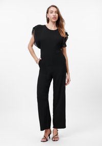 Body nero con maniche strutturate a volant, corpino aderente e pantaloni a gamba larga. La modella indossa tacchi con cinturino e ha le mani nelle tasche.