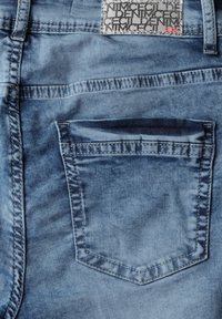 Tessuto denim blu con finitura sbiadita. Presenta una tasca posteriore e cuciture visibili. Etichetta con il nome del marchio in vita.