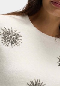 Bílý svetr s jemnou texturou, se stříbrně zdobenými designy slunečních paprsků na límci. Klasický kulatý límec.