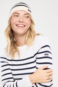 Jonge vrouw die glimlacht, gekleed in een wit en marineblauw gestreept trui en bijpassende muts, met haar armen over elkaar, en een beetje opzij kijkend.