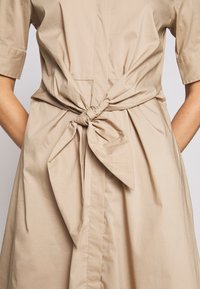 Beige bomullsklänning med korta ärmar, framknäppning och knytning i midjan. Slät textur med avslappnad passform.