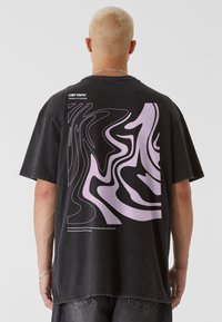 Černé oversized tričko s velkým fialovým abstraktním grafickým motivem na zádech, s textem pod designem v bílé barvě. Krátké rukávy, volný střih.