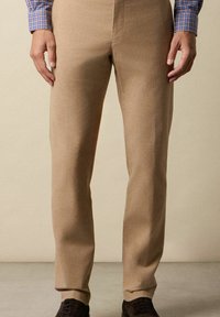 Beige Hosen im geraden Schnitt, aus strukturiertem Stoff, mit Gürtelschlaufen und Seitentaschen, kombiniert mit dunklen Schuhen.
