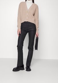 Beige stickad cardigan med V-ringning, tre knappar, kort passform, kombinerad med högmidjade svarta byxor och kraftiga svarta skor, håller i en svart clutch.