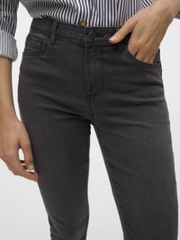 Sötétszürke farmert és kék-fehér csíkos gombos inget viselő személy, egyik kezével az elülső zsebében.