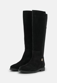 Bottes noires montantes en daim, présentant une texture lisse, un bout rond, un petit talon carré et un accent logo doré sur le côté.