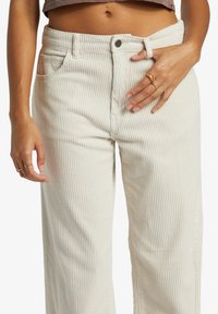 Pantalon large en velours côtelé crème avec des côtes verticales, design à cinq poches et fermeture par bouton. Tissu texturé avec un subtil éclat.