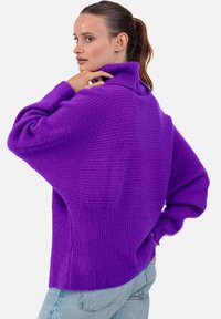 Pull en tricot côtelé violet avec un col montant et une coupe décontractée. Caractérisé par des manches surdimensionnées et un tissu texturé. Porté avec un jean bleu clair.