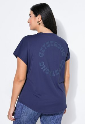 Pitkät tummahiuksinen nainen pukeutuu tummansiniseen paitaan, jossa on takana pyöreä "GET STRONG"-teksti, ja kuvioidut siniset leggingsit, katsoo vasemmalle.