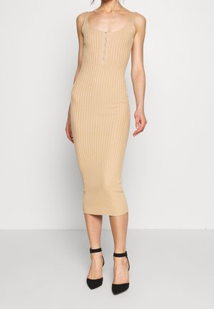 Woman wearing a fitted, ribbed beige midi dress with thin straps and buttons down the front, paired with black ankle-strap heels.