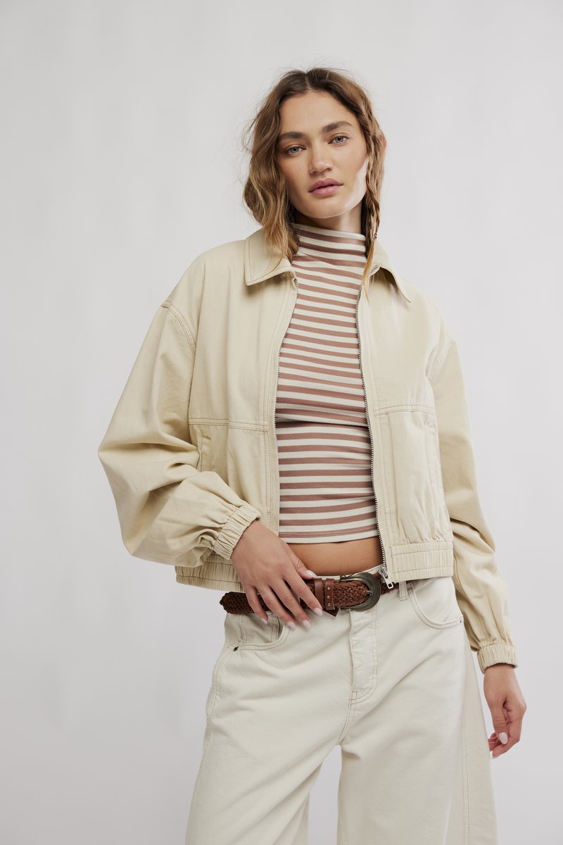 Beige kurze Jacke mit Reißverschluss vorne, elastischen Bündchen und schrägen Taschen, getragen über einem gestreiften, gerippten Shirt, kombiniert mit hellcremefarbenen Hosen und einem geflochtenen Gürtel.