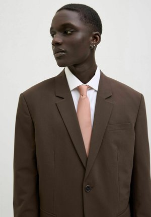 Costume marron avec un bouton unique, revers crantés et une poche poitrine ; associé à une chemise blanche et une cravate rose clair. Texture du tissu lisse.