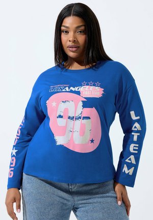 Kvinde iført en blå langærmet skjorte med pink og hvide tekster "Los Angeles First Team 96" og "LATEAM", kombineret med lyseblå jeans.