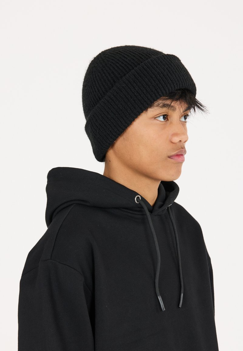 Svart stickad mössa med ribbat mönster, bärs något sned. Kombineras med en svart hoodie med dragkedja och avslappnad passform.